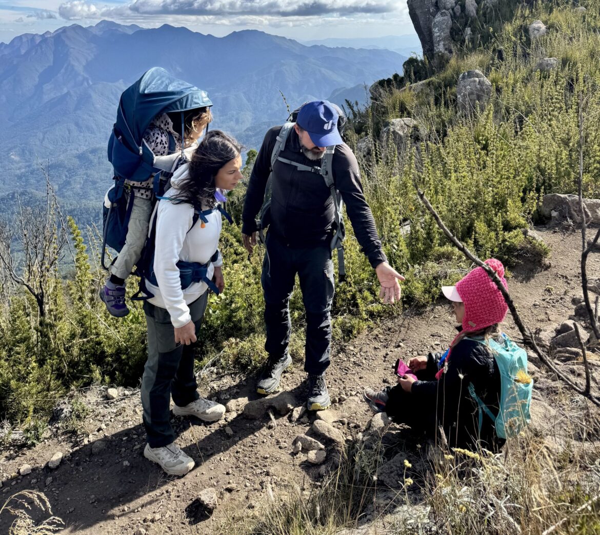 Familia parada em trilha de montanha observando o caminho, com crianca sentada em primeiro plano