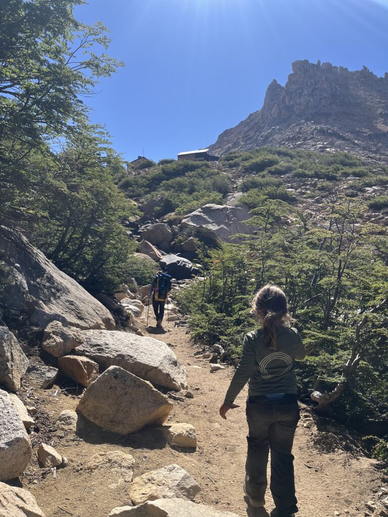 Pessoas caminhando em trilha rochosa em direção a um refúgio de montanha