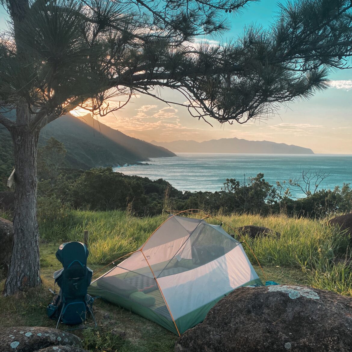 Barraca de acampamento montada sob uma arvore com vista para o mar ao entardecer