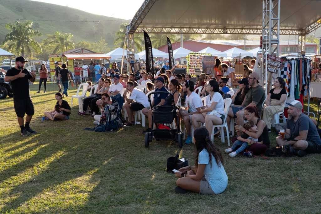 Fernando Barros falando para uma plateia sentada ao ar livre em evento outdoor com famílias e crianças